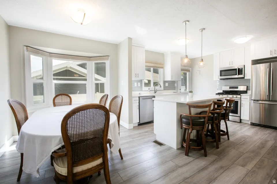 Open-concept kitchen and dining area featuring a bay window, wood-finish flooring, white cabinetry, a kitchen island with seating, and stainless steel appliances