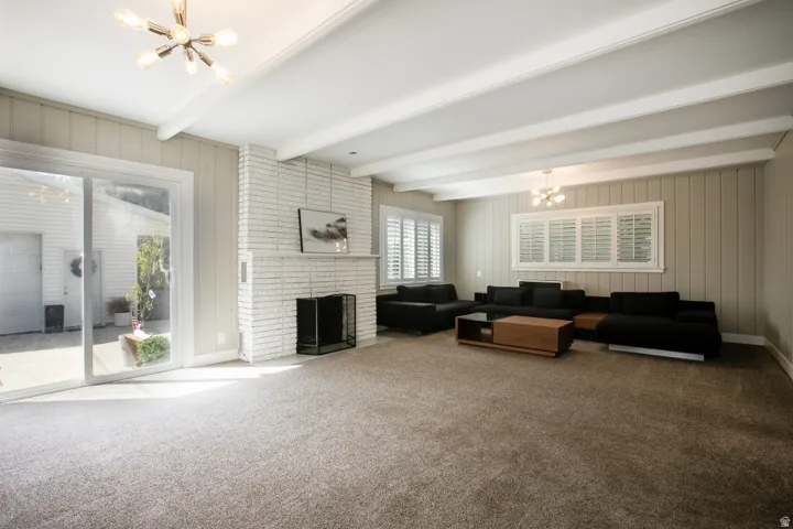 Spacious living area featuring exposed ceiling beams, a white brick fireplace, sliding glass doors, white plantation shutters, and a modern ceiling light fixture
