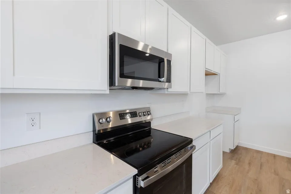 White shaker-style cabinetry, stainless steel microwave, black electric range, light-toned countertops, and wood-finish flooring