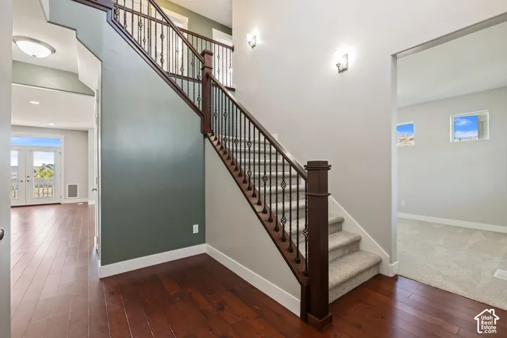 Wood-finish flooring, staircase with carpeted treads, dark wood railing with wrought iron balusters, recessed wall sconces, and double French doors