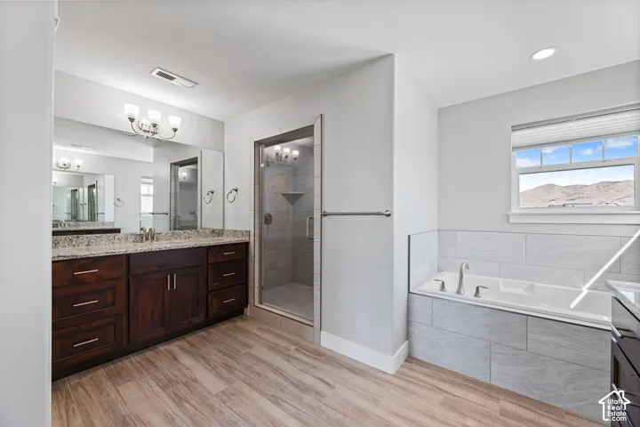 Spacious bathroom featuring a dual vanity with a granite countertop, wood-finish flooring, a glass-enclosed shower, and a built-in soaking tub with tiled surround