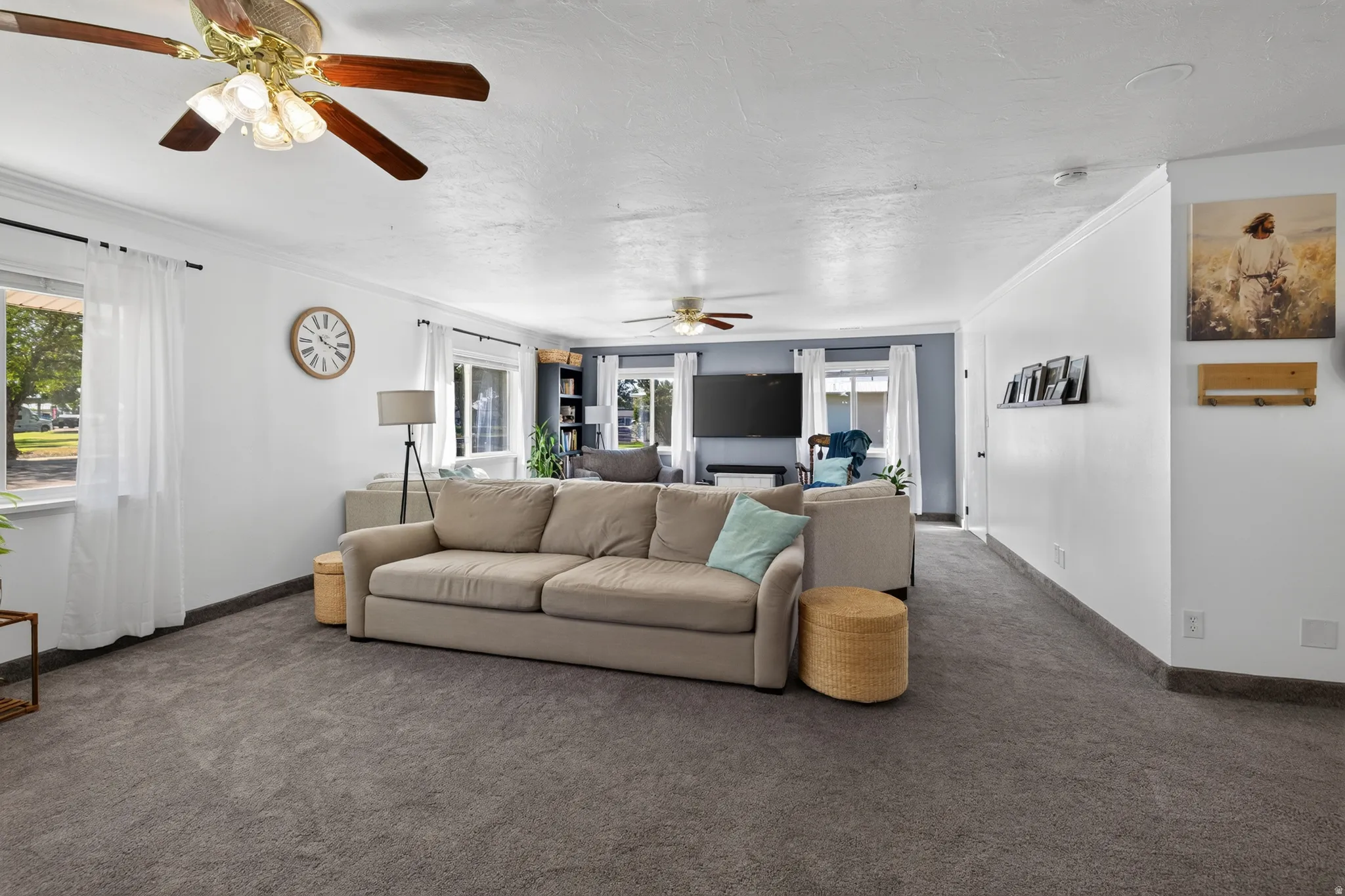 Expansive living area with gray carpeting, multiple windows, and two ceiling fans with light fixtures