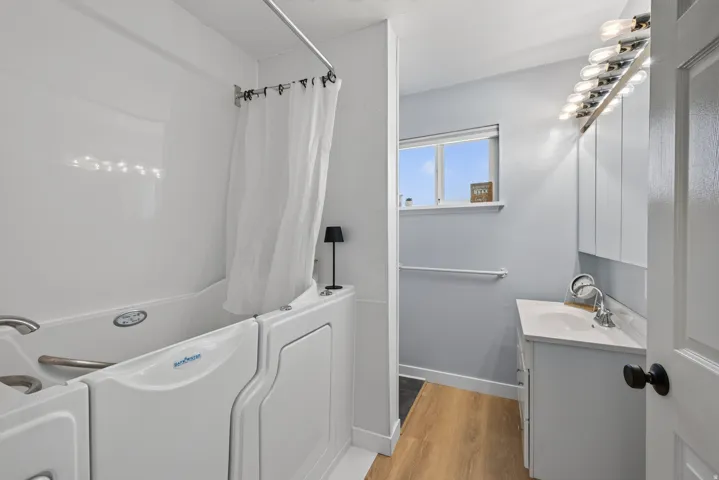 Walk-in tub with grab bars, wood-finish flooring, single vanity with integrated sink, and upper cabinetry