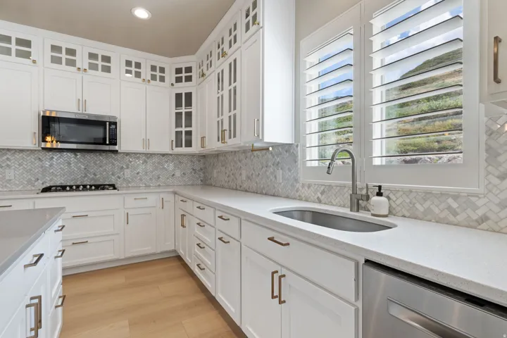 White cabinetry with brushed bronze hardware, herringbone tile backsplash, integrated stainless steel appliances, light-toned wood-finish flooring, and plantation shutters