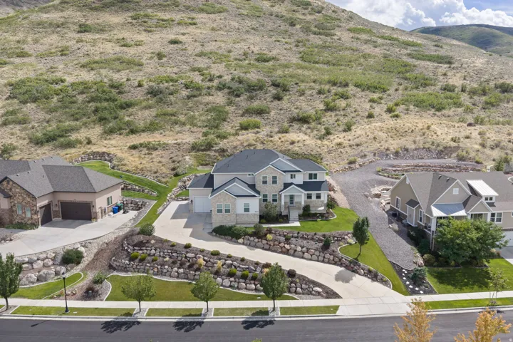 Two-story residence with a gray shingle roof and light-colored siding, featuring a curved concrete driveway and tiered landscaping with natural rock elements