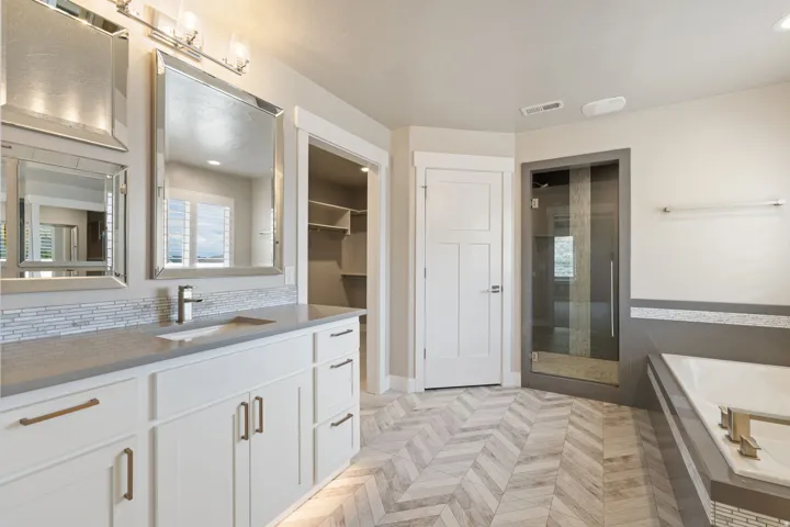 Dual vanity with integrated sinks, herringbone-patterned tile flooring, frameless glass shower enclosure, built-in bathtub, and an open closet with shelving