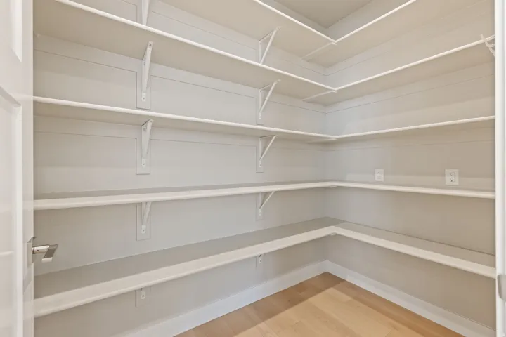 Expansive walk-in pantry with light wood-finish flooring, white built-in shelving, multiple wall outlets, and a white paneled door