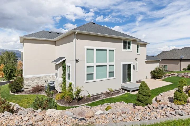 Stucco exterior with stone accent, dark shingle roof, multiple windows, sliding glass door, and a concrete patio