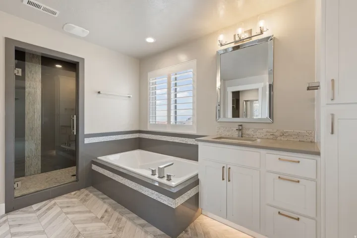 Spacious bathroom featuring a glass-enclosed shower, built-in soaking tub with tile surround, white vanity with integrated sink and stone countertop, and herringbone-patterned floor tile