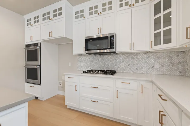 White cabinetry with bronze hardware, stainless steel double ovens, a built-in microwave, a gas cooktop, and a herringbone tile backsplash