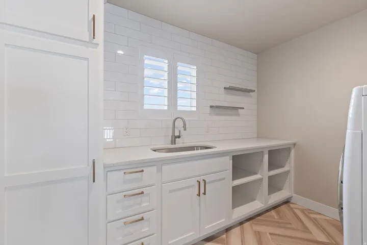 White cabinetry with bronze hardware, subway tile backsplash, integrated sink with gooseneck faucet, light-toned wood-finish flooring, and open shelving