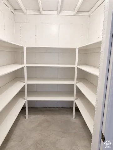 Walk-in storage area featuring white painted concrete block walls, multi-level tiered shelving, and exposed ceiling joists