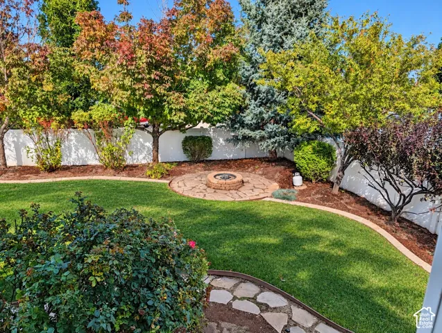 Backyard featuring a stone-paver fire pit, manicured lawn, curved concrete curbing, mature trees, and perimeter vinyl fencing