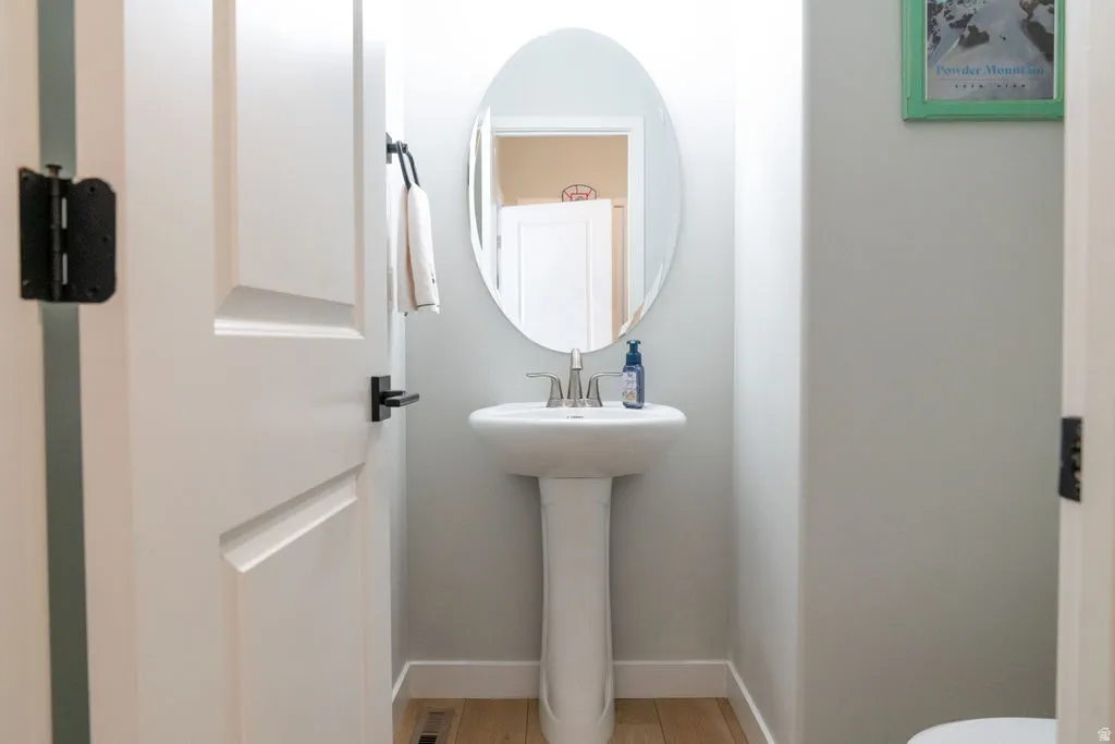 Pedestal sink with stainless steel faucet, oval mirror, and light wood-finish flooring