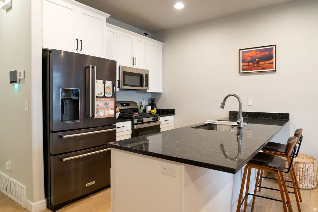 Kitchen featuring white shaker cabinetry, stainless steel appliances, a center island with a sink, black stone countertops, and light wood-finish flooring