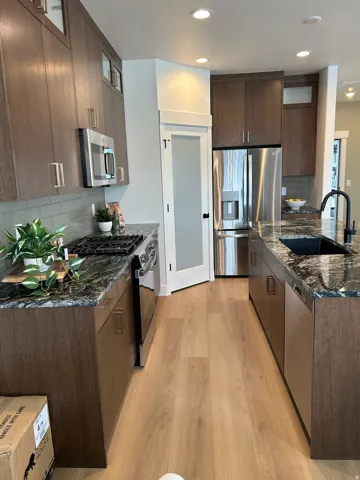Kitchen featuring wood-finish flooring, dark stone countertops, stainless steel appliances, dark wood-tone cabinetry, and a black sink with a matte black faucet