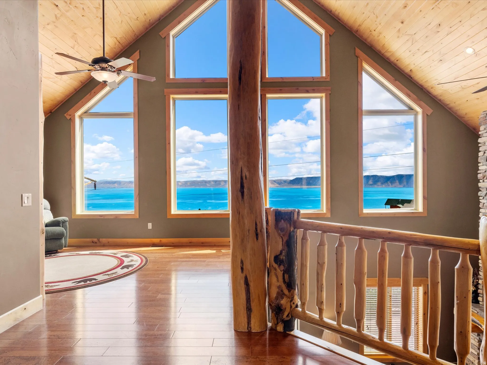 Expansive windows with trapezoid transoms, natural wood-finish vaulted ceilings, wood-finish flooring, a wood-finish ceiling fan, and a rustic wood railing with balusters