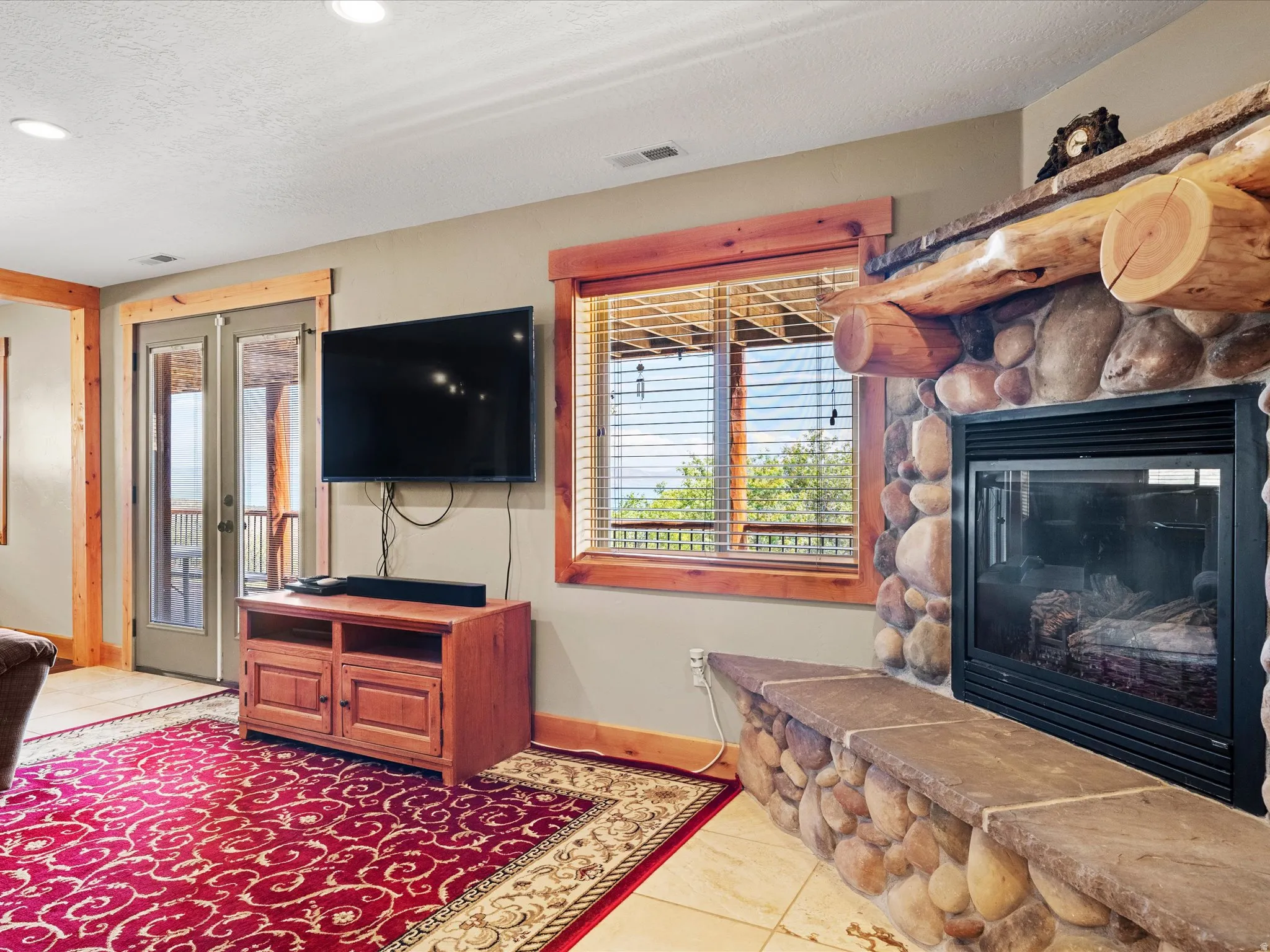 Stone fireplace with log mantel and hearth, recessed lighting, wood-trimmed double glass doors, and tiled flooring