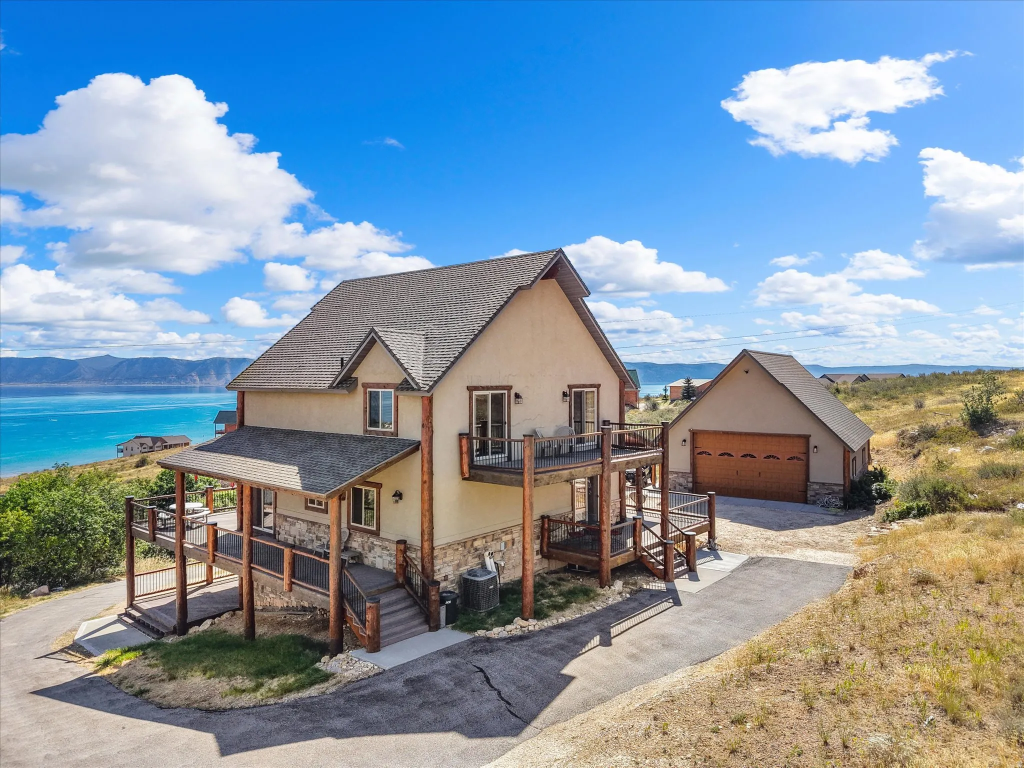 Multi-level exterior with stucco and stone accents, featuring a multi-tier deck with wood railings, a detached garage with an arched door, and a prominent lake backdrop