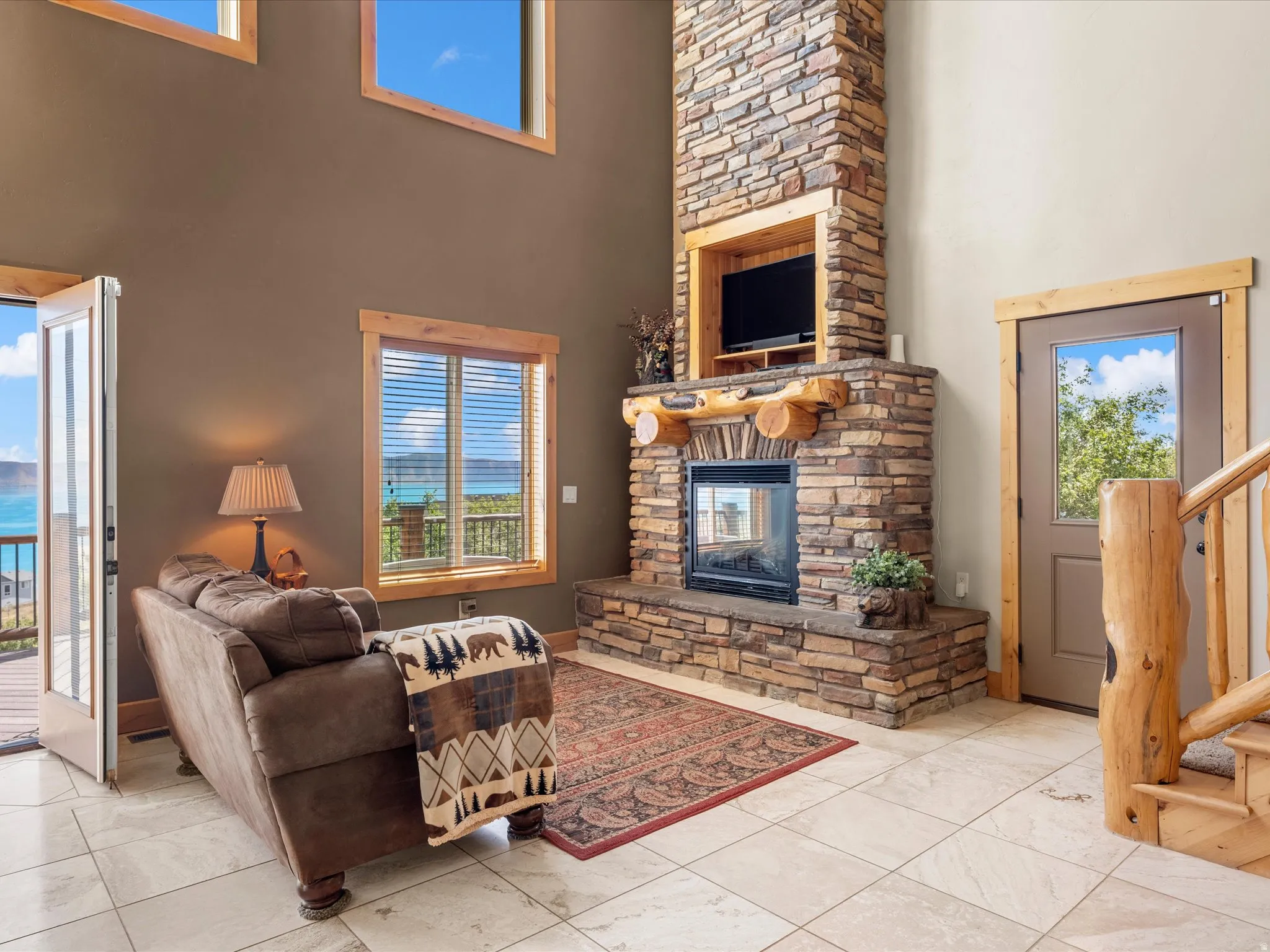 Stone fireplace with wood mantel, high ceilings, large windows with natural scenery, light-toned tile flooring, and wood-framed doors