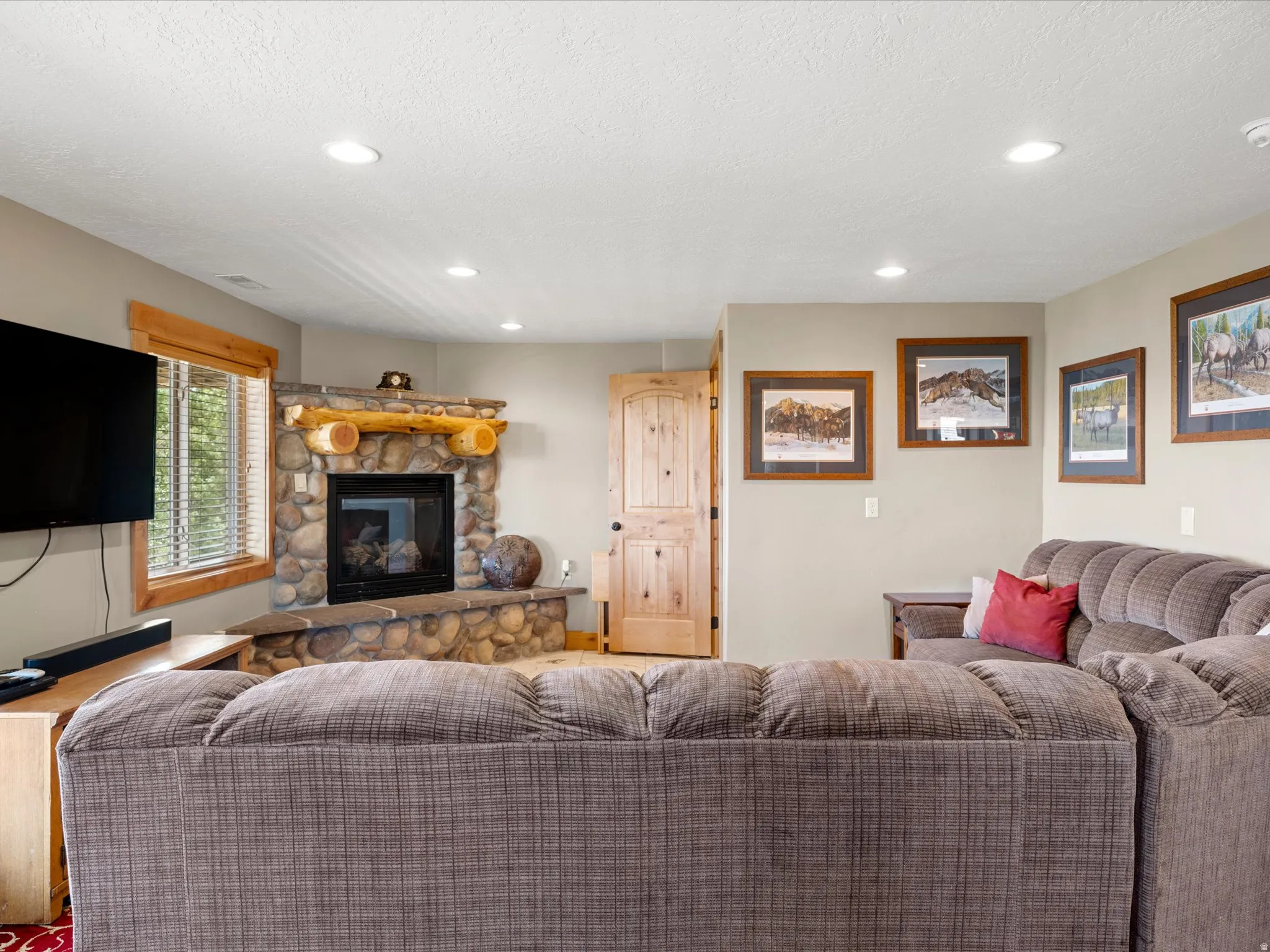 Stone-faced fireplace with a log mantel, recessed lighting, wood-finish window trim, and a paneled wood door