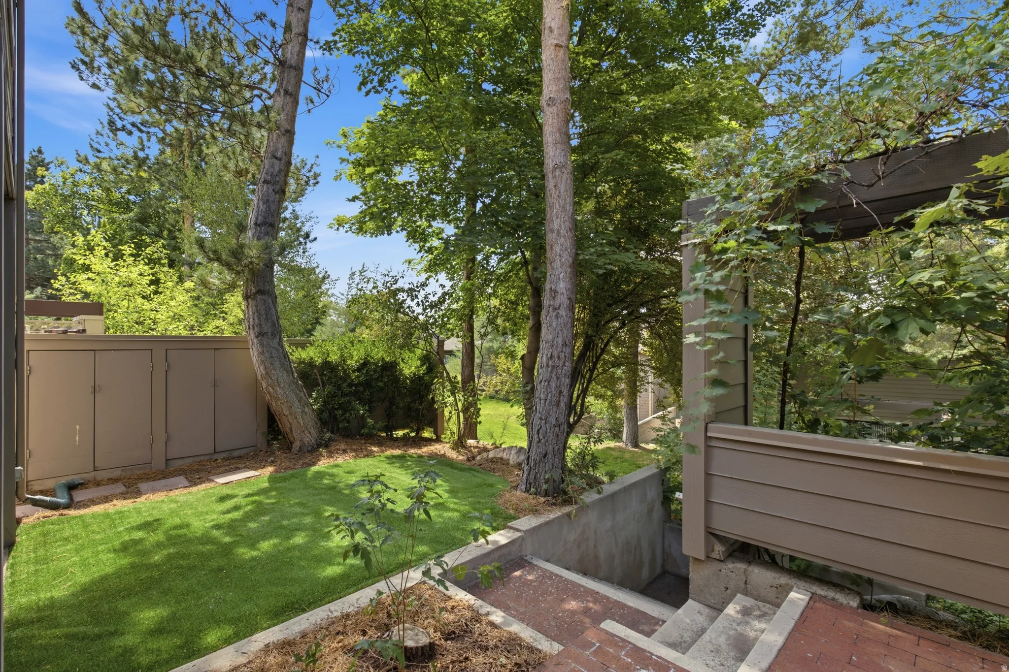 Manicured lawn area with mature trees, a built-in storage unit, concrete retaining walls, and brick-paved steps leading downwards
