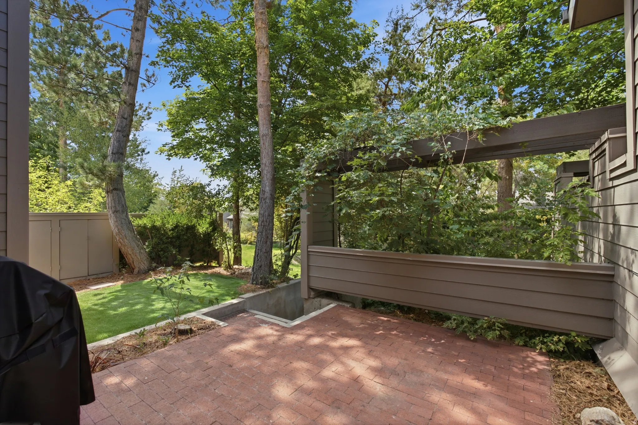 Brick paver patio with a wood pergola structure, surrounded by mature trees and a manicured lawn