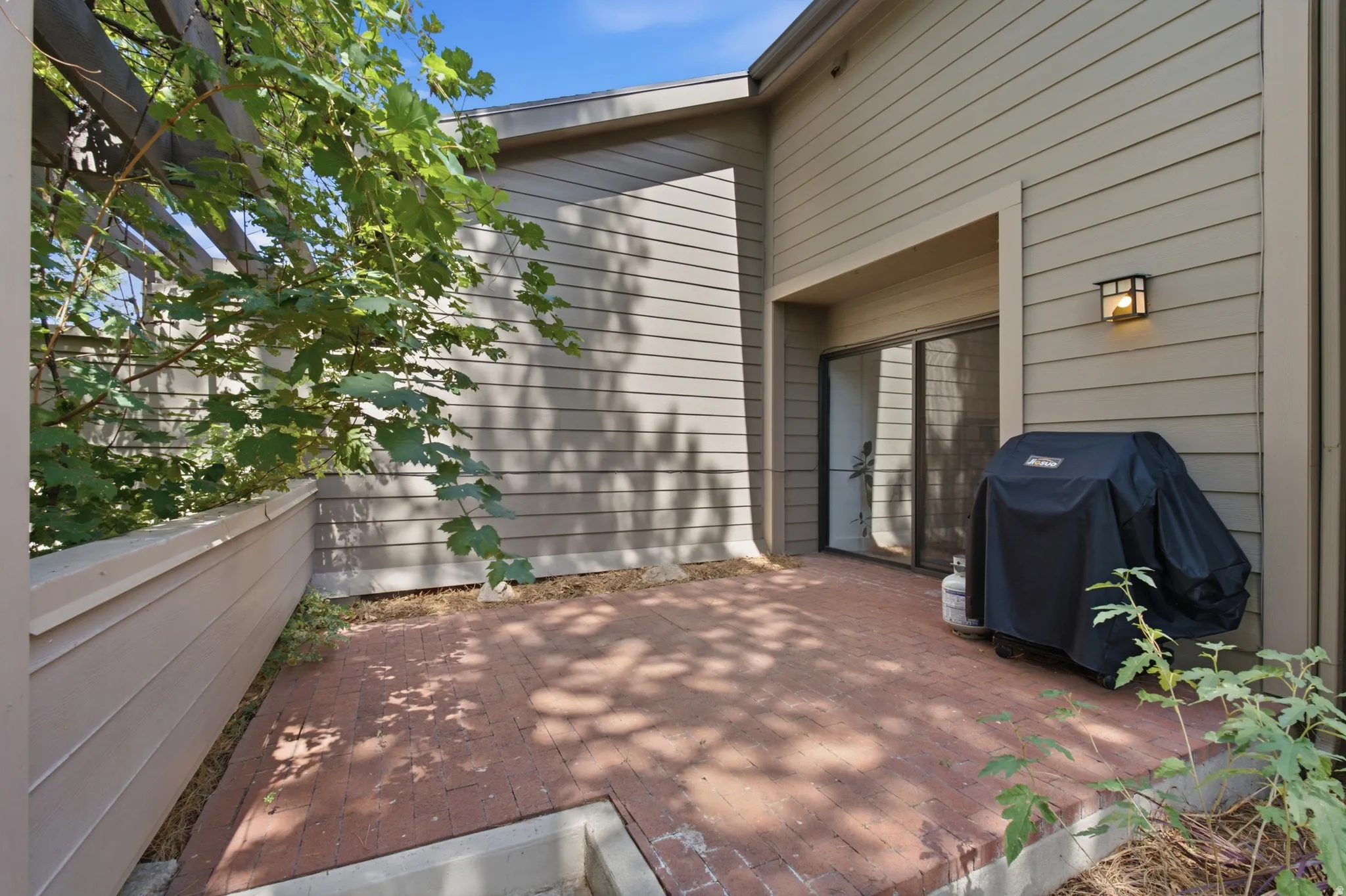 Private patio featuring a brick paver surface, wood privacy fencing, a wood trellis with natural foliage, and exterior wall lighting