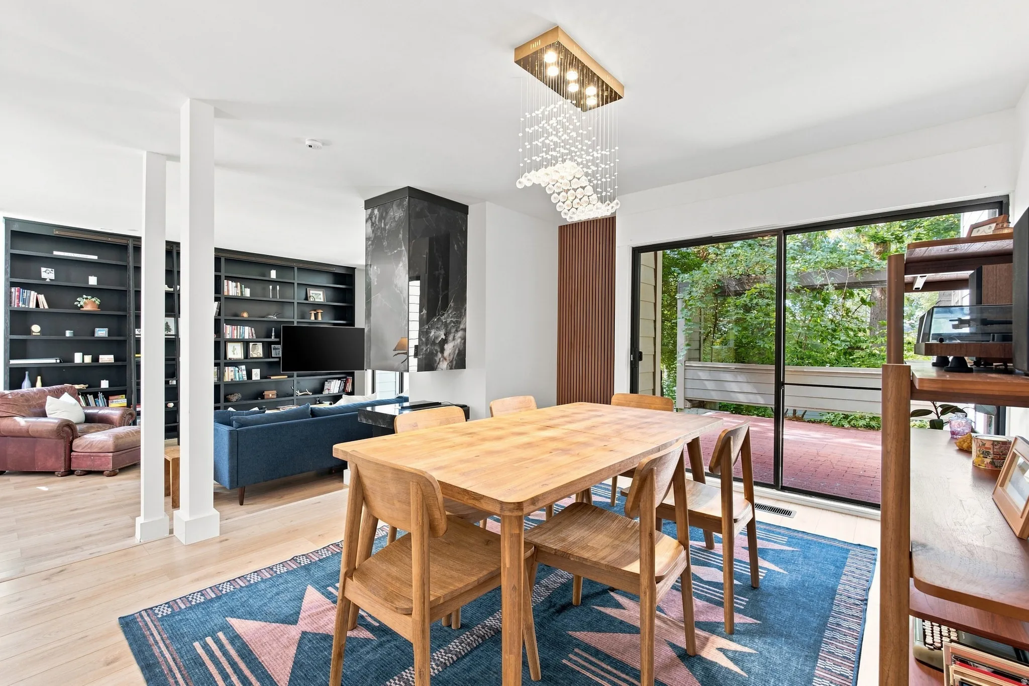 Open-concept living area featuring a natural wood dining table with seating for four, wide-plank wood-finish flooring throughout, and abundant natural light from expansive glass sliding doors