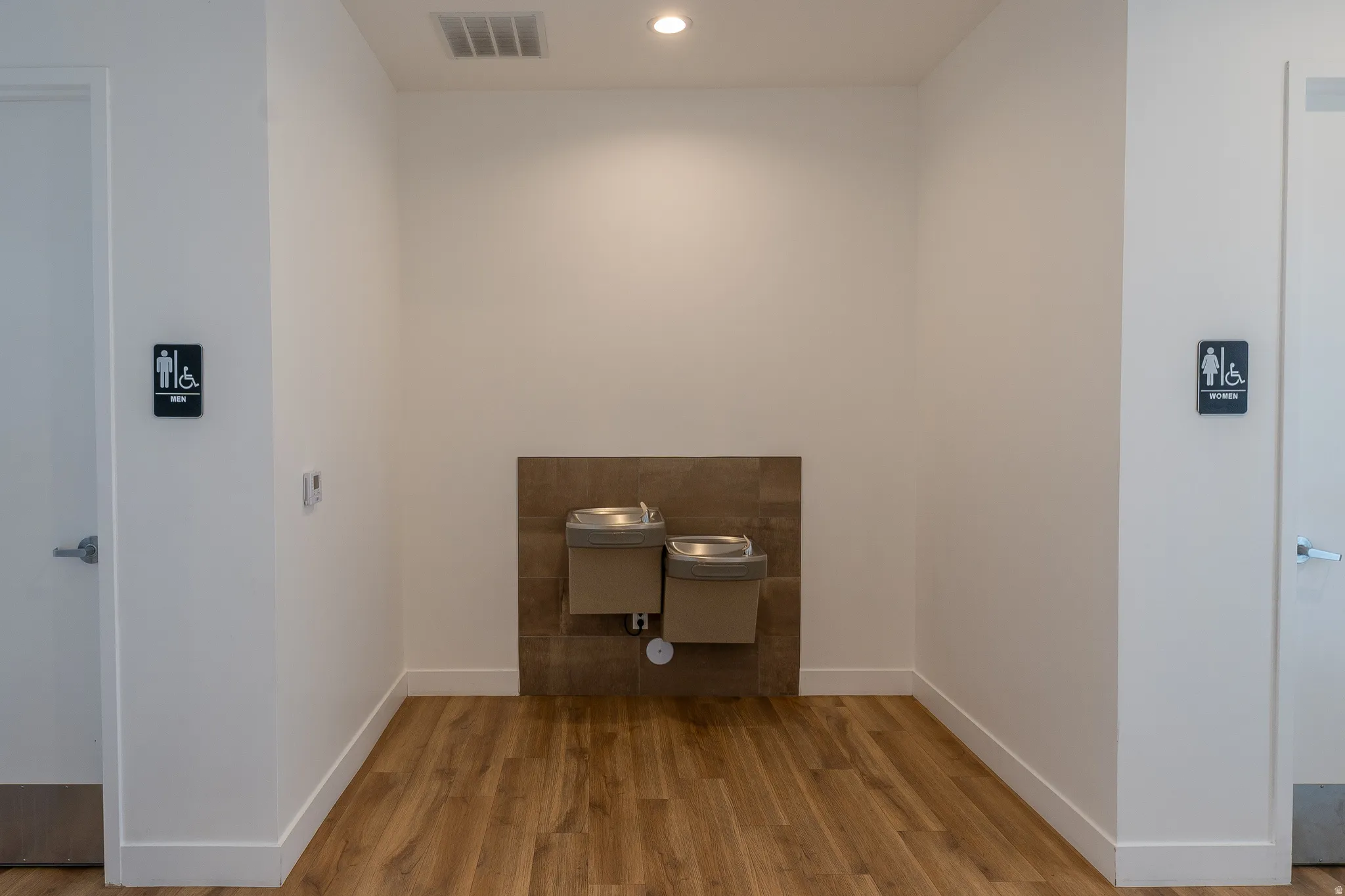 Wood-finish flooring, two stainless steel water fountains, recessed ceiling lighting, and white baseboards