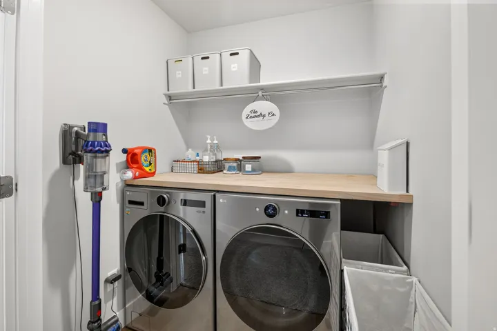 Full-sized laundry appliances with a wood-finish countertop, white wall shelving, and light gray walls