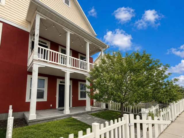 Two-story exterior with a red lower facade and light-colored siding, featuring a covered porch and upper balcony with white railings, a concrete walkway, and a white picket fence