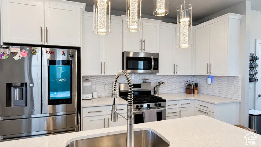 White shaker cabinetry, stainless steel appliances, patterned tile backsplash, light-toned countertops, and contemporary pendant lighting