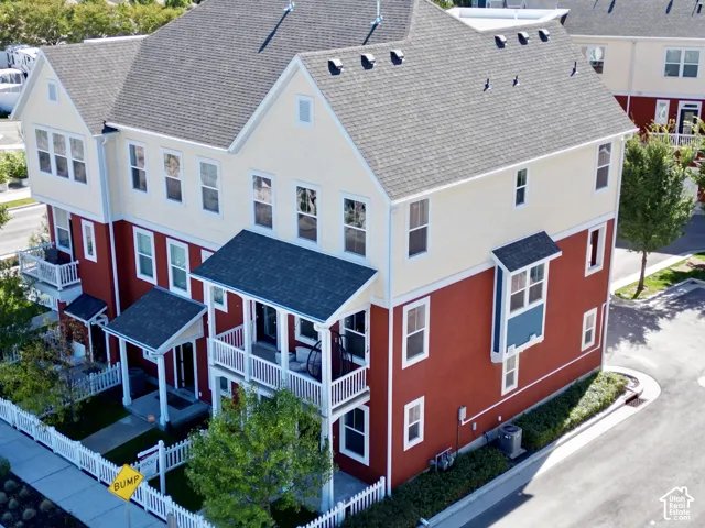 Three-story building with a multi-textured exterior featuring red lower-level cladding and cream upper-level siding, accented by dual-level balconies with white railings, a multi-gabled roof, and a white picket fence perimeter
