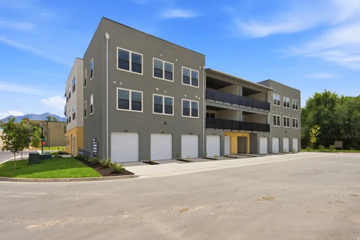 Modern multi-unit residential building featuring clean-lined architecture with a taupe exterior, expansive glazing across three stories, and ground-level garage parking with white overhead doors