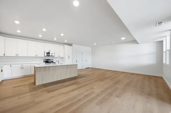 Open-concept kitchen and living area featuring white cabinetry with stainless steel appliances, a large center island with wood-finish detailing, recessed ceiling lighting, and wide-plank wood-finish flooring throughout the space