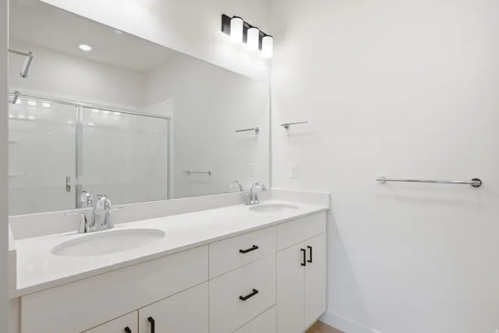 Dual undermount sinks with chrome faucets, white vanity with matte black hardware, large mirror, and overhead black and white vanity lighting