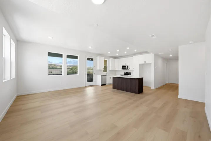 Expansive open-concept room featuring light wood-finish flooring, recessed lighting, and a kitchen with white cabinetry, stainless steel appliances, and a dark wood island with a light-colored countertop