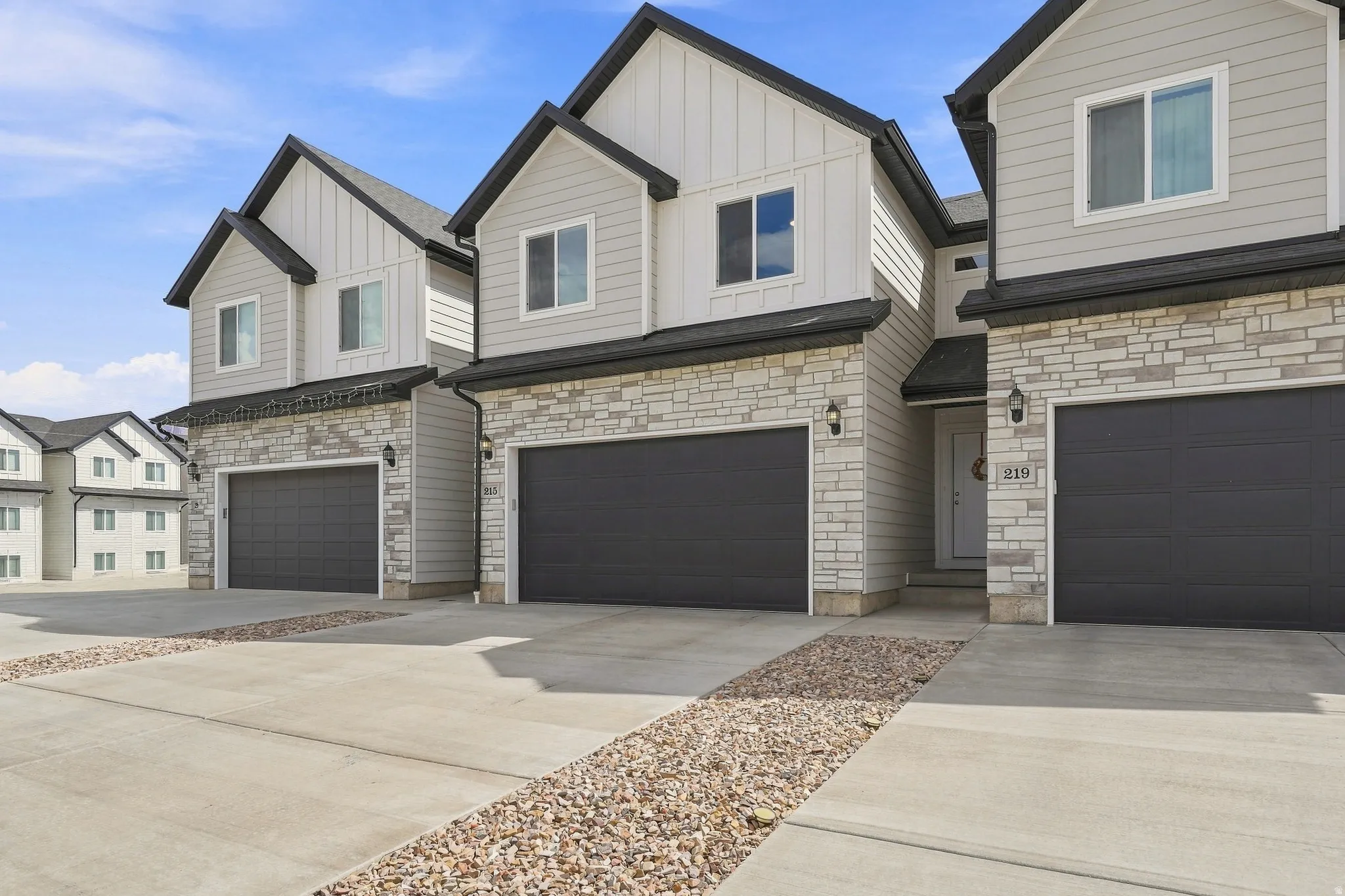 Modern townhome exterior featuring light-colored stone veneer, dark gray garage doors, and a concrete driveway with decorative stone accents
