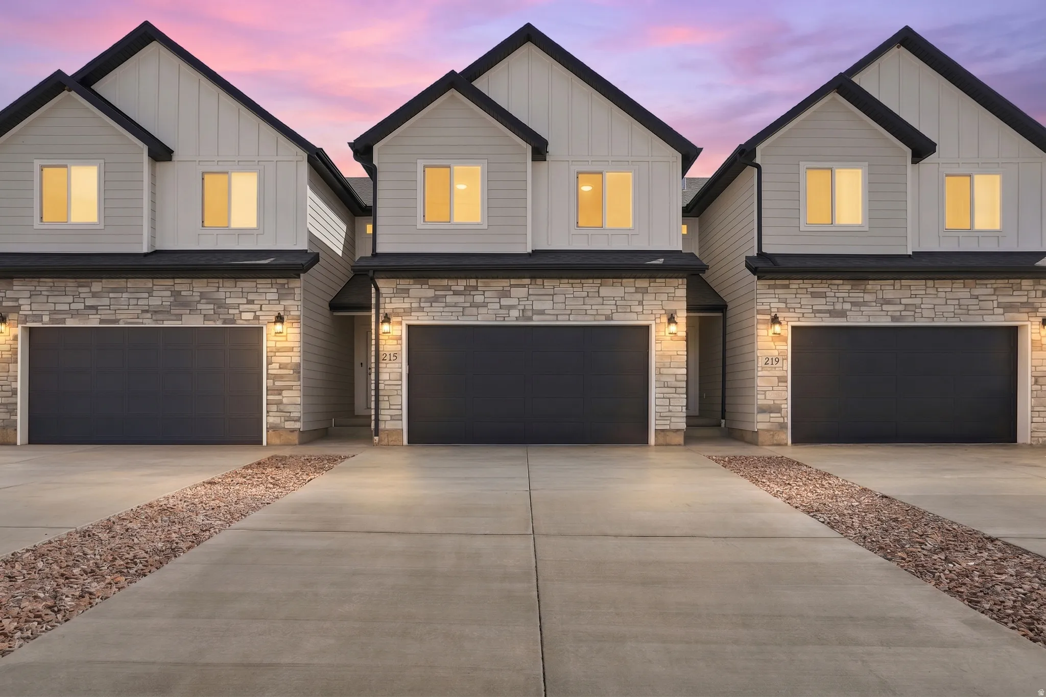 Modern exterior with stone accents and board and batten siding, featuring dark garage doors and concrete driveways with decorative rock strips