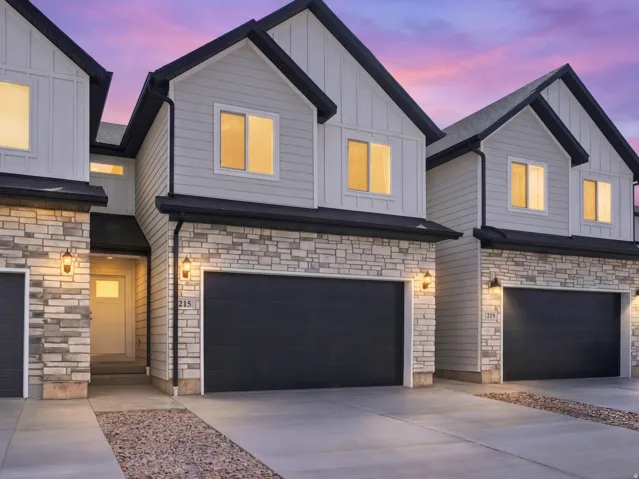 Modern townhome architecture featuring light-colored siding, stone facade accents, dark garage doors, and exterior wall-mounted lighting