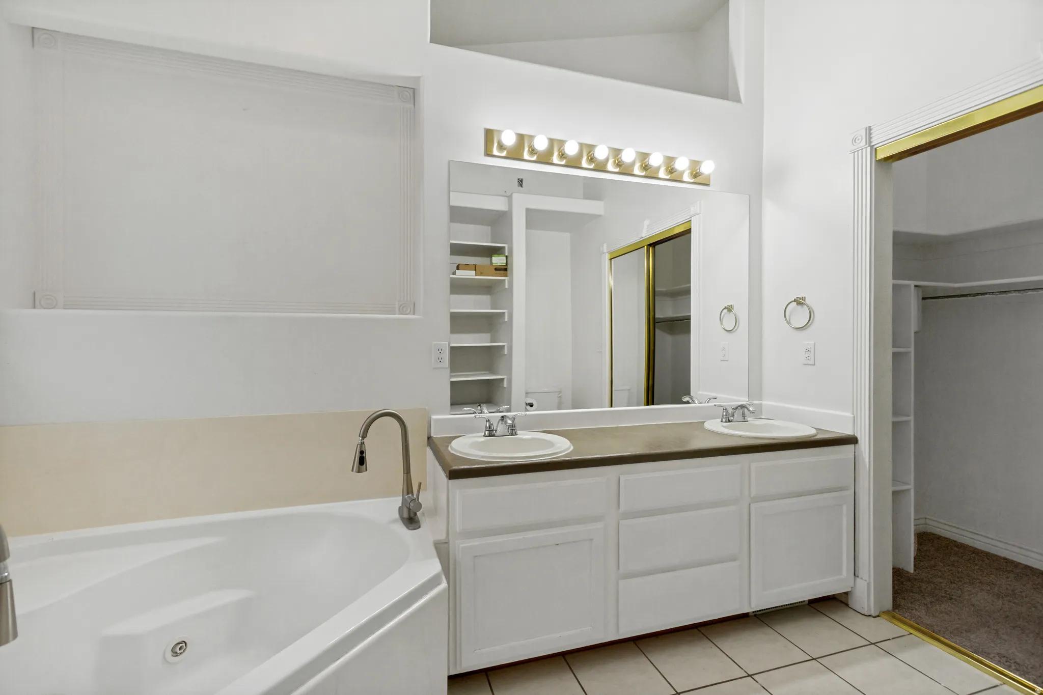 Spa-inspired primary bathroom featuring a soaking tub with deck-mounted faucet, dual vanity with wood-finish countertop and ample storage, illuminated mirror with brass-finish lighting, tile flooring, and brass-finish hardware throughout