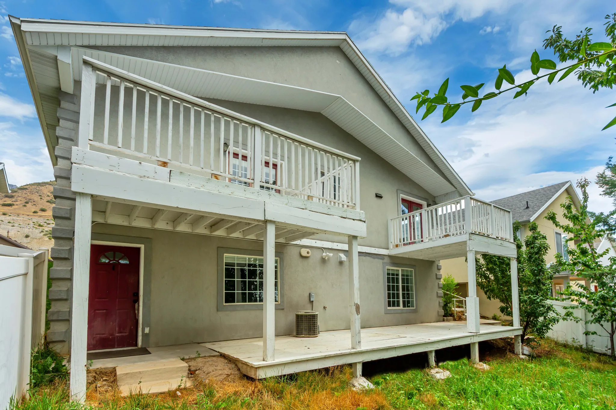 Charming two-story home featuring a covered front porch with white railings, a spacious ground-level deck ideal for outdoor entertaining, and lush landscaping with mature greenery