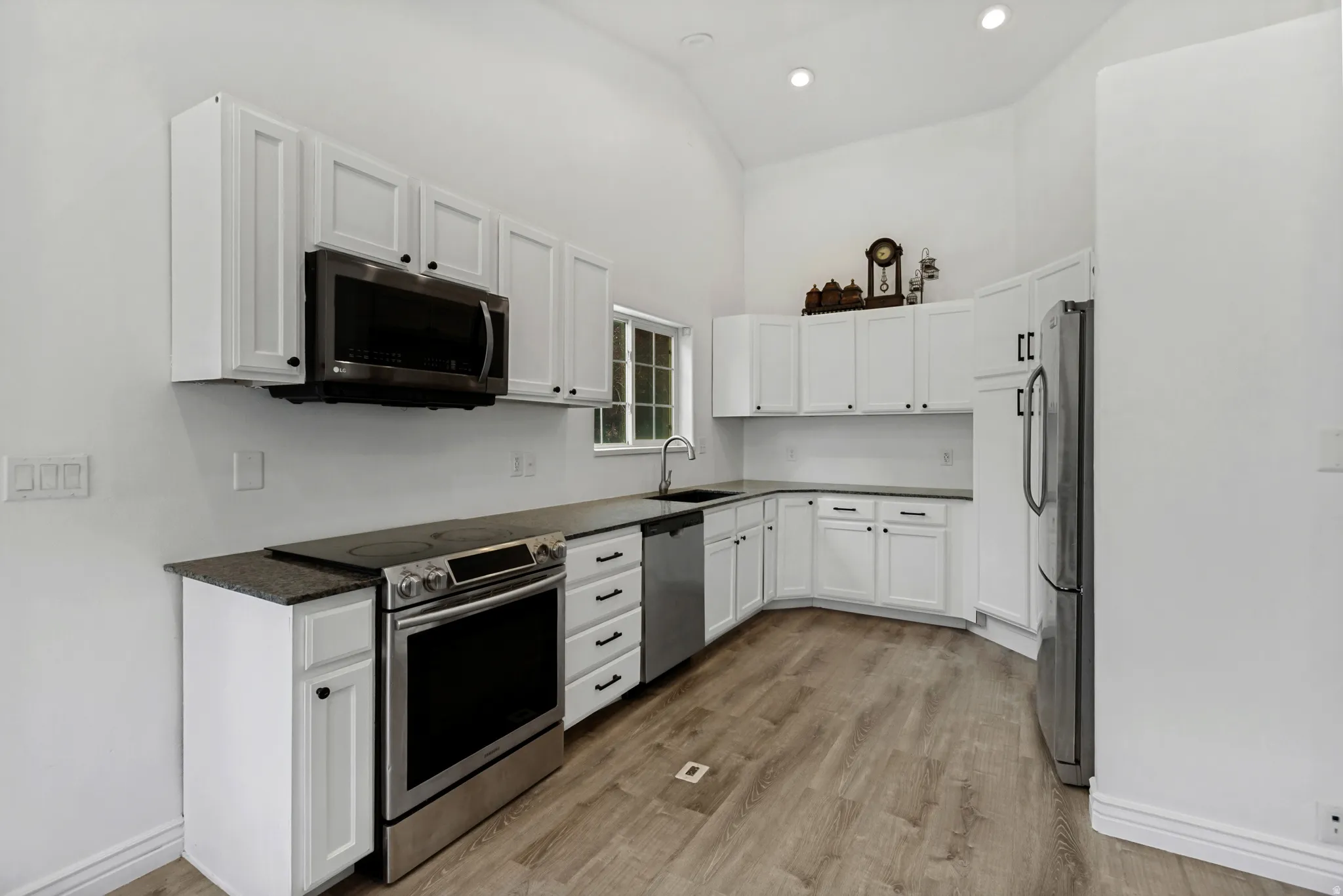 Kitchen featuring white shaker cabinetry, stainless steel appliances, dark countertops, wood-finish flooring, and a vaulted ceiling