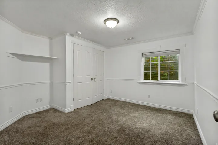 Built-in double closet with panel doors, recessed window with blinds, wall-mounted shelving, overhead lighting, and neutral carpeting