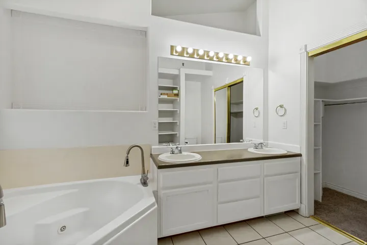 Spa-inspired primary bathroom featuring a soaking tub with deck-mounted faucet, dual vanity with wood-finish countertop and ample storage, illuminated mirror with brass-finish lighting, tile flooring, and brass-finish hardware throughout
