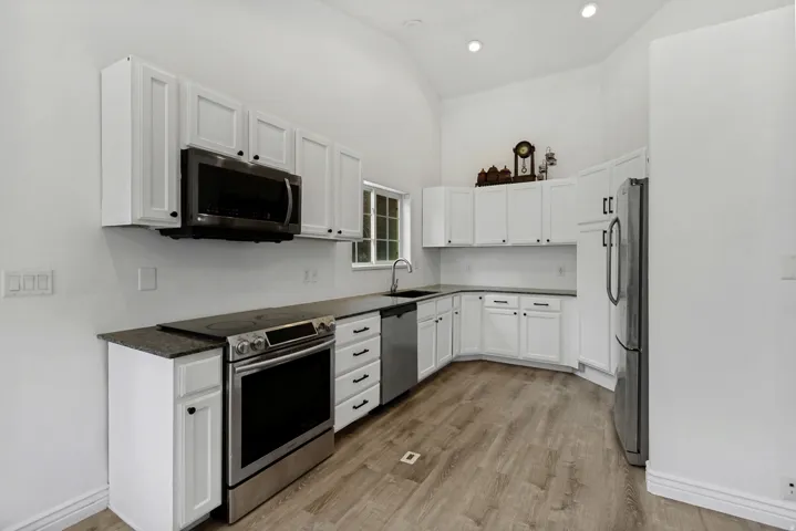 Kitchen featuring white shaker cabinetry, stainless steel appliances, dark countertops, wood-finish flooring, and a vaulted ceiling