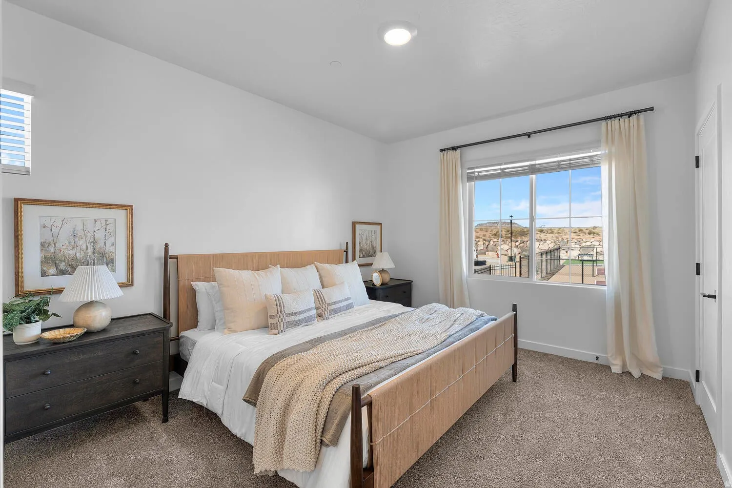 Bright primary bedroom featuring a wood-frame bed, dual nightstands with dark finishes, carpet flooring, recessed ceiling lighting, and expansive windows with custom drapery offering panoramic views of the surrounding landscape