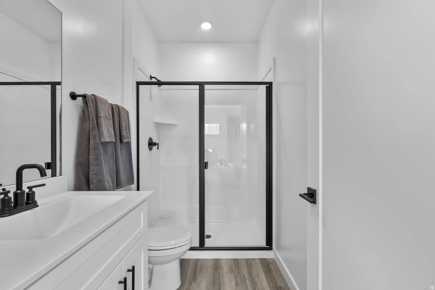 Modern bathroom featuring a white vanity with black fixtures, a glass-enclosed shower with black framing, wood-finish flooring, and a recessed ceiling light