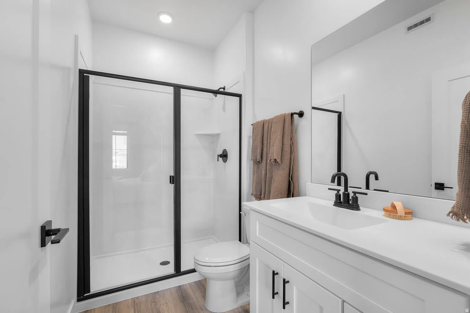 Bathroom featuring a white vanity with integrated sink, black fixtures, a glass-enclosed shower, wood-finish flooring, and a large wall-mounted mirror